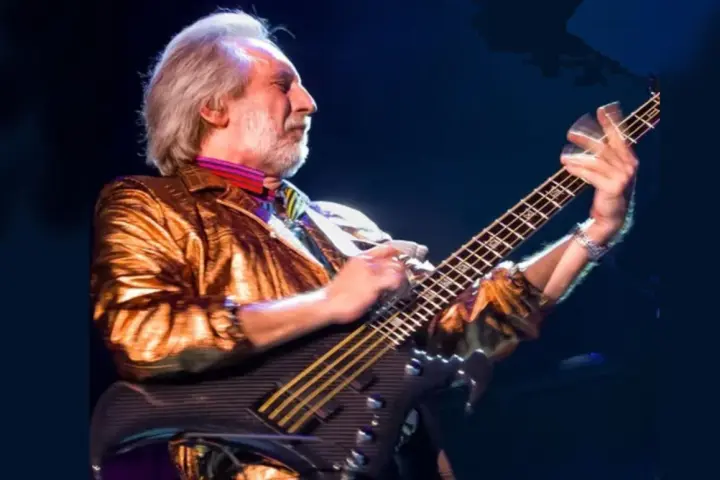 Ein Mann mit grauem Haar und Bart spielt Bassgitarre. Er trägt eine glänzende goldene Jacke und bunte Krawatte. Er ist der beste Bassist der Welt.