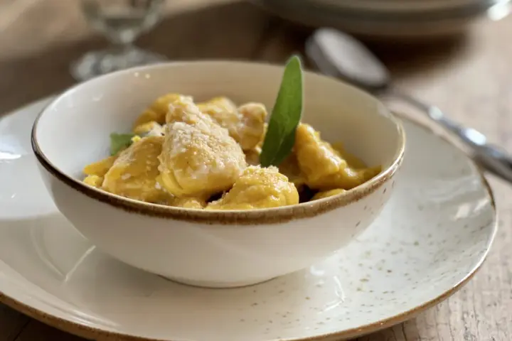 Gnocchi di zucca dorati e soffici, conditi con parmigiano e una foglia di salvia, serviti in una ciotola bianca su un piatto maculato.