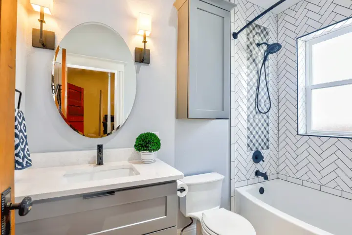 Une petite salle de bain moderne avec baignoire, lavabo gris, miroir rond et carrelage chevrons. Une belle idée pour optimiser l'espace.