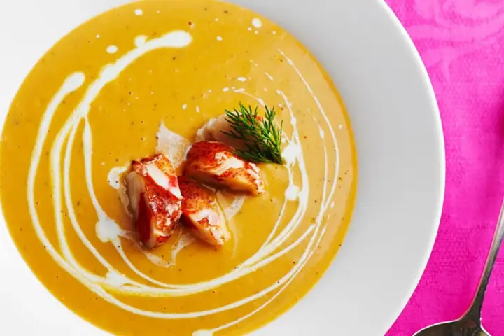 Un cremoso bisque de langosta, adornado con trozos de carne de langosta y un toque de eneldo, servido en un plato blanco sobre un mantel rosa vibrante.