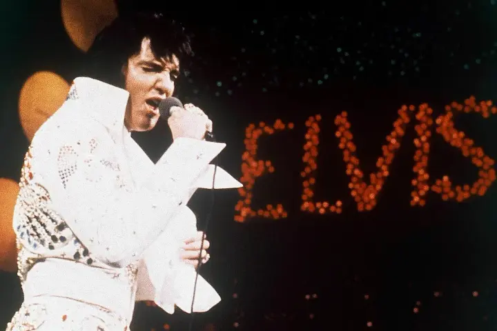 Elvis Presley singt in seinem berühmten weißen Anzug in Las Vegas, während sein Name in roten Lichtern erstrahlt.