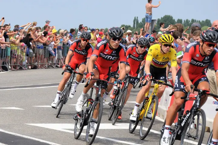 Cyclists in red BMC jerseys race, with one in yellow, as spectators cheer. A perfect scene to watch Tour de France live!