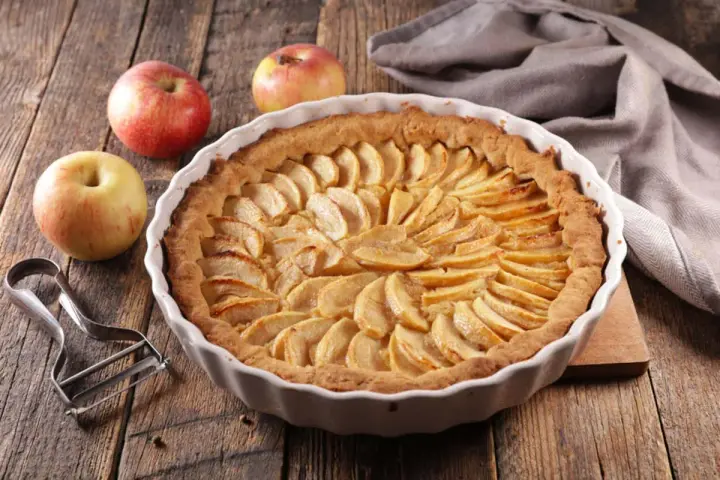 Torta di mele umida e soffice, con mele fresche e un pelapatate su un tavolo di legno rustico.