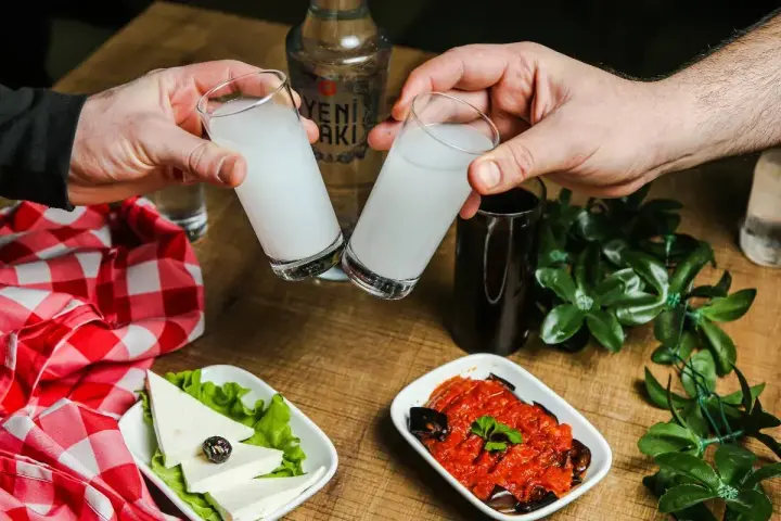 Dwie szklanki z rakı, tradycyjnym tureckim alkoholem, unoszą się do toastu. Obok ser, sałata, sos pomidorowy z bakłażanem i butelka rakı. Idealne połączenie na wieczór.