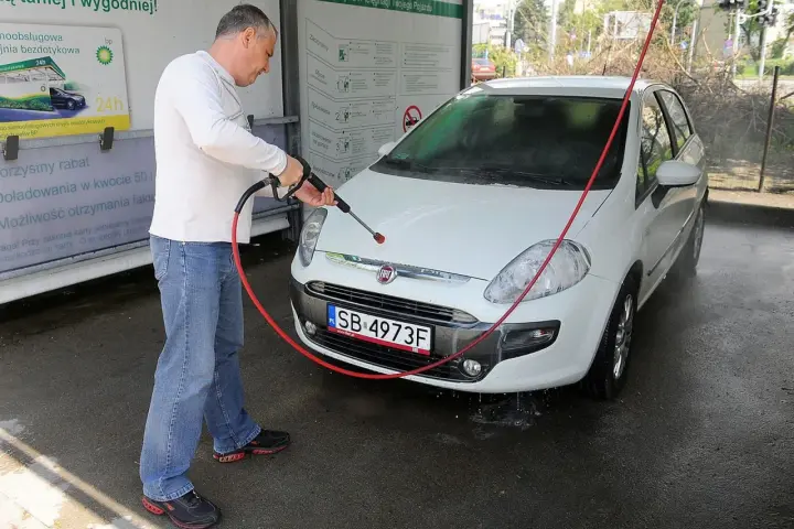 Mężczyzna myje białe auto na myjni. Na tablicy informacyjnej widnieje napis "Możliwość otrzymania faktury", sugerujący, że na myjni można płacić kartą.