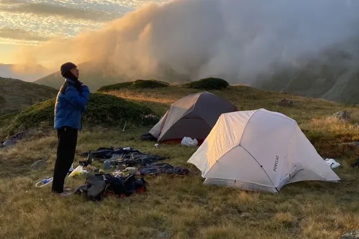 Une personne se tient près de tentes dans une aire de bivouac, profitant du lever du soleil sur les montagnes.