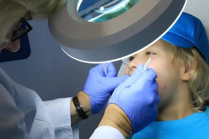 Dentysta w rękawiczkach bada usta dziecka pod lampą powiększającą.