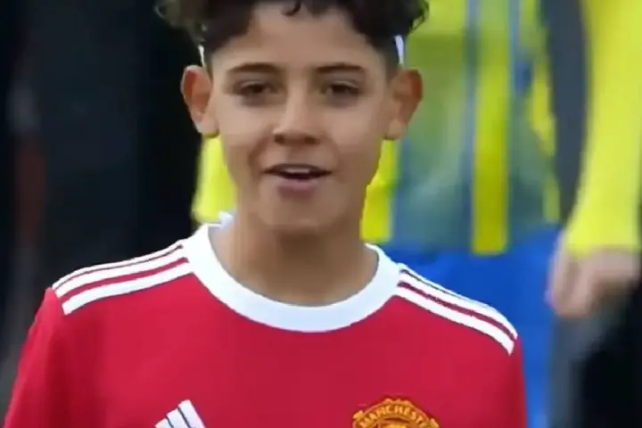 Młody piłkarz w czerwonej koszulce Manchester United, przypominający syna Cristiano Ronaldo. Jego wiek sugeruje, że to początki kariery.
