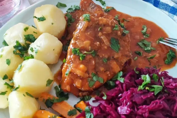 Pyszna szynka w sosie własnym, podana z ziemniakami, marchewką i czerwoną kapustą. Danie posypane jest świeżą natką pietruszki.