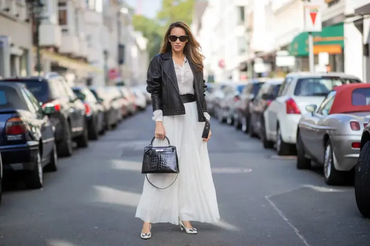 Elegancka kobieta w białej, plisowanej spódnicy i czarnej ramonesce. Idealne buty do plisowanej spódnicy to te, które widzisz: białe czółenka z czarną kokardką.