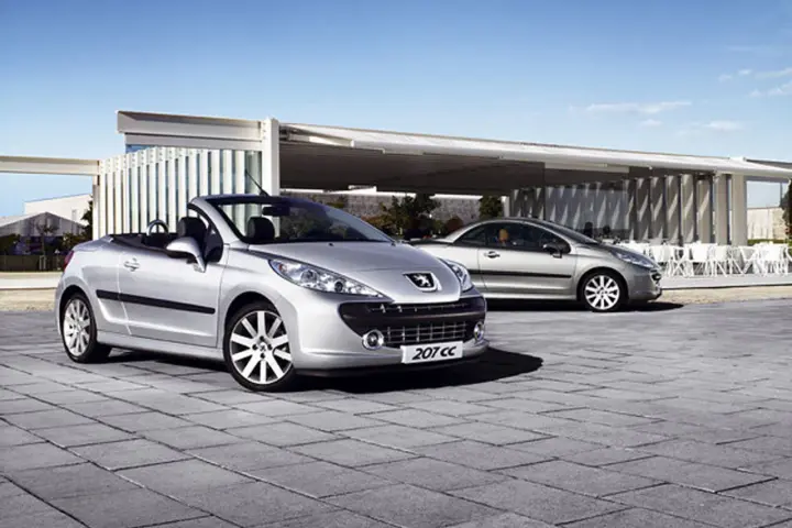 Två silverfärgade Peugeot 207 CC cabriolet står parkerade på en stenlagd uteplats framför en modern byggnad.