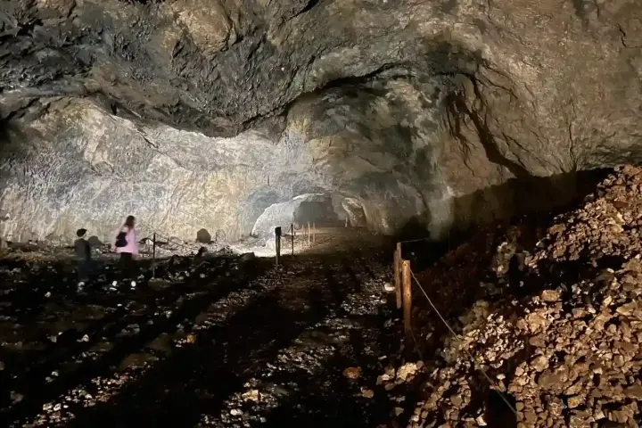 Wnętrze jaskini ciemnej, z kamienistym dnem i skalnymi ścianami. W oddali widać dwie osoby zwiedzające.