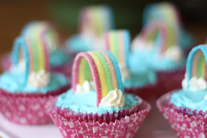 Sju cupcakes med ljusblå regnbågs frosting och godisregnbågar. Rosa muffinsformar med mönster.