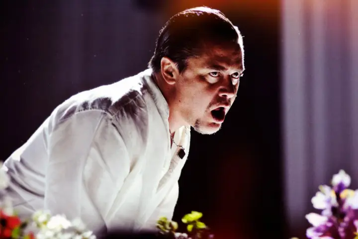 Mike Patton heute, mit weit aufgerissenem Mund und intensivem Blick, trägt ein weißes Hemd. Blumen im Vordergrund.