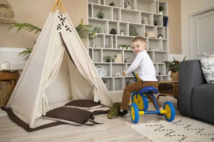 Un petit garçon sur un tricycle bleu et jaune s'amuse dans un espace de jeux, près d'une tente tipi et d'un canapé. Ce centre de vacances et de loisirs est idéal pour les enfants.