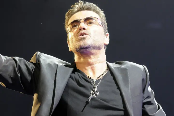 George Michael w okularach, w czarnej marynarce i z naszyjnikiem, śpiewający swoje niezapomniane utwory.