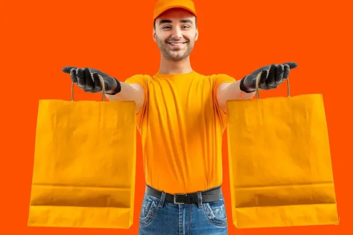 Un rider di Glovo azienda, con guanti neri e maglia gialla, porge due borse arancioni.
