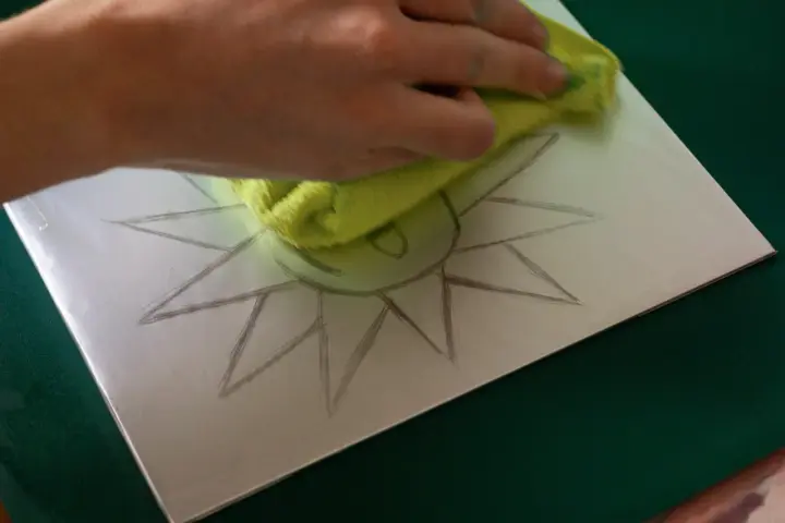 Mano limpiando una superficie metálica con un paño verde, sobre la cual se dibuja un sol. Preparando una litografía casera.