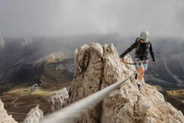 Kobieta w kasku i uprzęży pokonuje via ferrata, balansując na linie nad skalistym zboczem. W tle rozciąga się malownicza dolina.