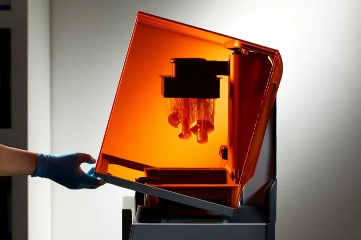 A gloved hand opens the orange lid of a stereolithography 3D printing machine, revealing intricate parts being built layer by layer.