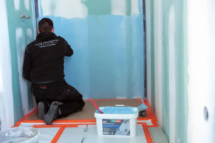 Un ouvrier applique un système d'étanchéité liquide bleu sur les murs d'une douche. Un seau de produit est visible au premier plan.