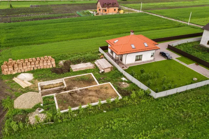 Działka 1000m2 wymiary: rozpoczęta budowa domu, gotowy fundament, sterta cegieł, drewno, żwir, obok stoi dom z pomarańczowym dachem i samochód.