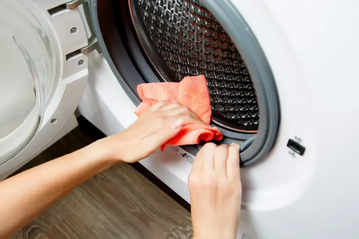 Hands cleaning the rubber seal of a washing machine with an orange cloth. This is part of how to clean washing machine.
