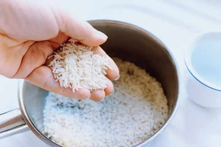 Mano sosteniendo arroz para calcular el tiempo cocción arroz perfecto. Olla con arroz y taza de agua al lado.