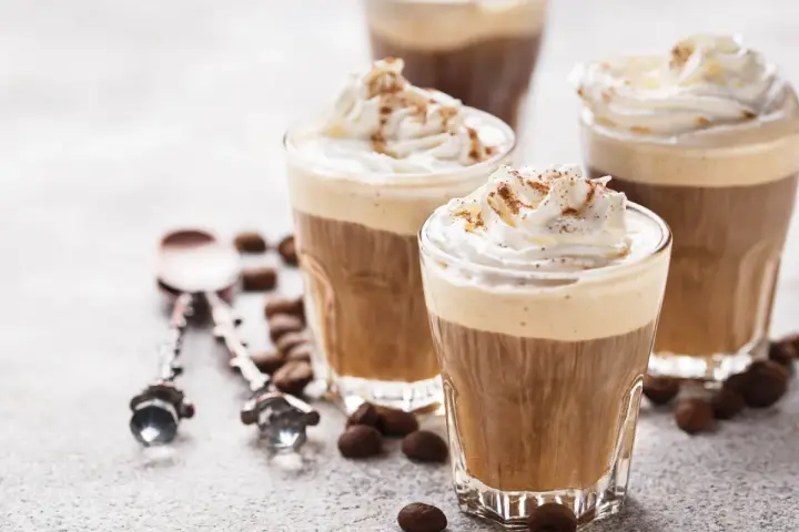 Tre bicchieri di deliziosa crema al caffè, guarniti con panna montata e cacao. Chicchi di caffè e cucchiaini decorati completano la scena.
