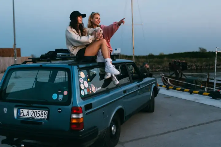 Dwie kobiety siedzą na dachu niebieskiego kombi przy nabrzeżu, jedna wskazuje palcem w dal.