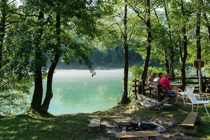 Spokojny poranek na polu kempingowym nad jeziorem. Mgła unosi się nad wodą, a przy stole siedzi kilku mężczyzn.