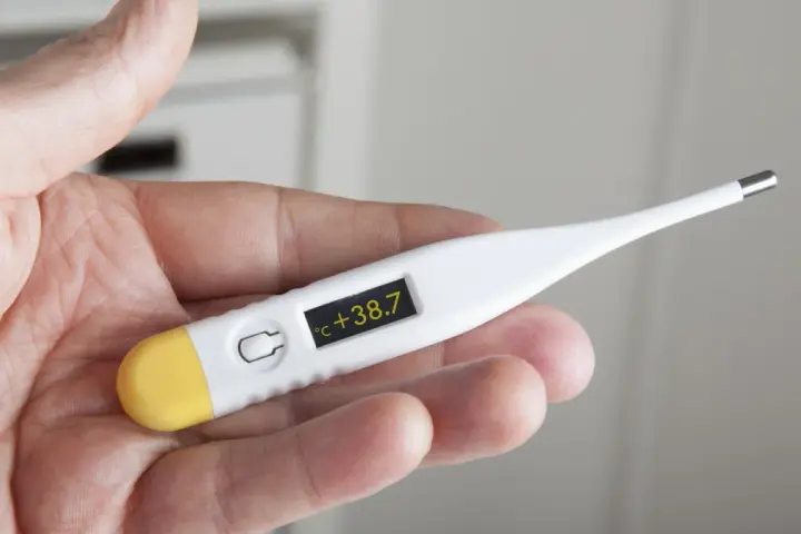Dłoń trzyma termometr wskazujący 38.7°C. Czas na działanie, jak zbić gorączkę.