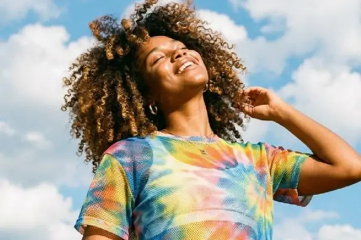 Une femme souriante aux cheveux bouclés, sous un ciel bleu nuageux. Elle porte un t-shirt tie-dye coloré. Une belle image pour apprendre comment rendre une photo flou.