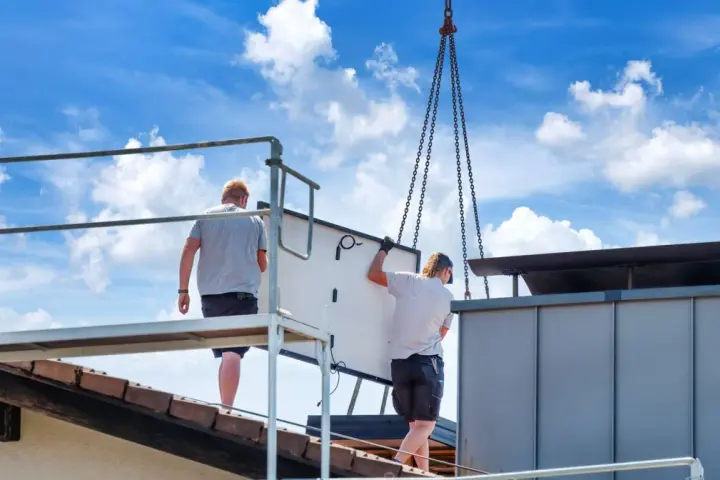Dwaj pracownicy montują panel fotowoltaiczny na dachu. Ciężka praca i ryzyko wypadku to negatywne skutki fotowoltaiki, które często się pomija.