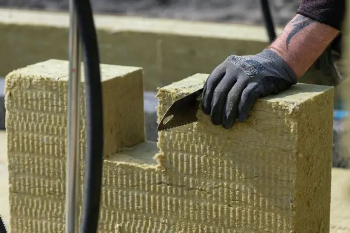 Ręka w rękawicy tnie piłą wełnę skalną 10. Materiał izolacyjny jest cięty na budowie.