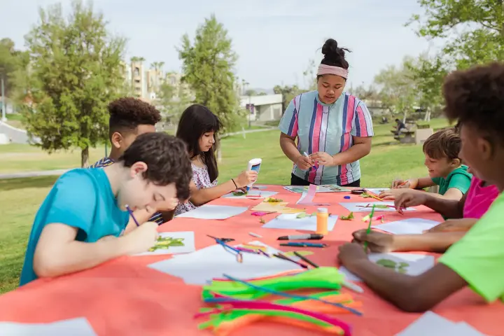Des enfants créent des œuvres d'art en plein air autour d'une table recouverte d'une nappe orange.