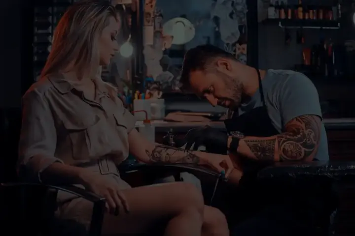 Artysta tworzy tatuaż opaska na nodze kobiety. W tle widać studio tatuażu.