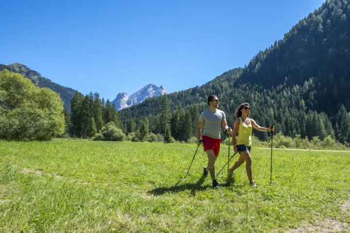 Coppia fa nordic walking in un prato verde con montagne sullo sfondo, ideale per canazei escursioni facili.