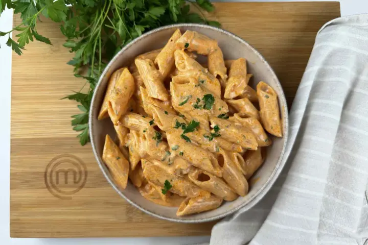 Penne alla vodka cremose e invitanti, guarnite con prezzemolo fresco, servite in una ciotola su un tagliere di legno con il logo MasterChef.
