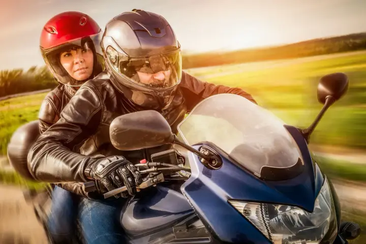 Coppia in moto, con lui alla guida e lei dietro, entrambi con caschi e giacche di pelle. Un'immagine che evoca la libertà di come guidare una moto.