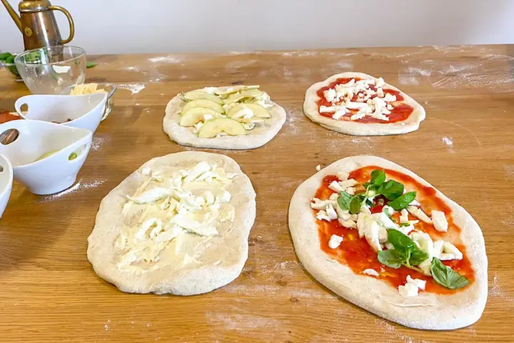 Vier vorbereitete Pinsa-Teiglinge liegen auf einem Holztisch. Einer mit Apfelscheiben, einer mit Tomatensoße und Mozzarella, einer nur mit Mozzarella und einer mit Tomatensoße, Mozzarella und Basilikum.