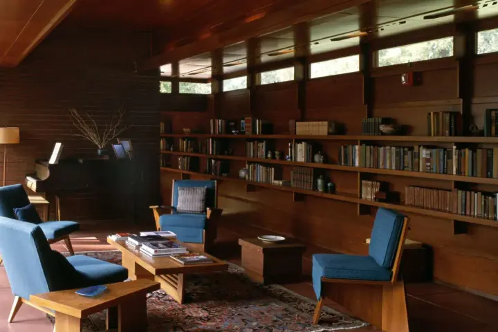 A cozy living room with blue chairs, a piano, and bookshelves. The clerestory windows above let in natural light, illuminating the warm wood interior.