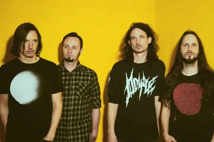 Quatre hommes aux cheveux longs posent devant un fond jaune vif. Leurs t-shirts portent des graphismes distinctifs, évoquant l'énergie brute du magma et la puissance de Gojira.
