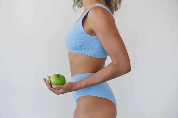Mujer en bikini azul sostiene una manzana verde, símbolo de una dieta rápida para bajar de peso y un estilo de vida saludable.