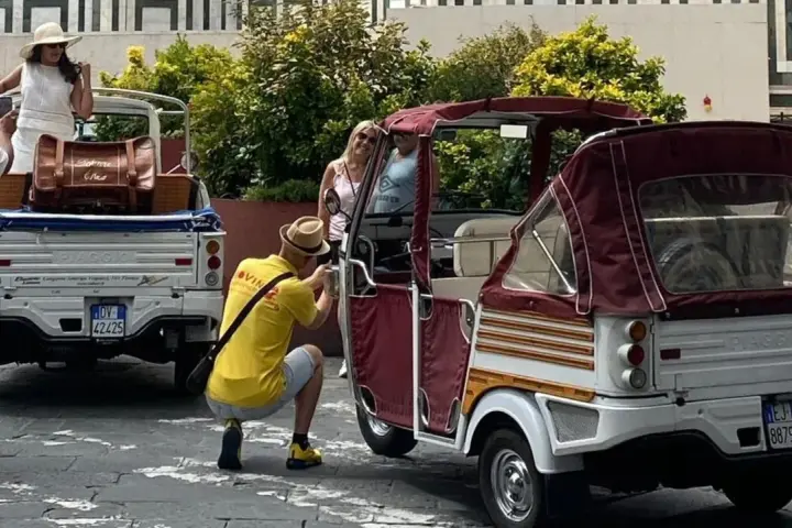 Turisti scattano foto a veicoli atipici, un Piaggio Ape e un tuk-tuk, in una strada soleggiata.