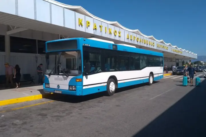 Niebiesko-biały autobus na tle budynku lotniska z żółtym napisem.