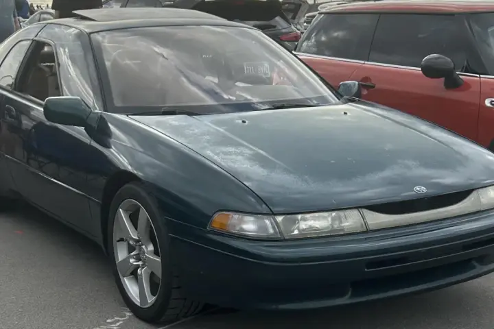 Subaru SVX w ciemnozielonym kolorze, z charakterystycznymi oknami w stylu "glass-canopy", zaparkowany obok czerwonego samochodu.