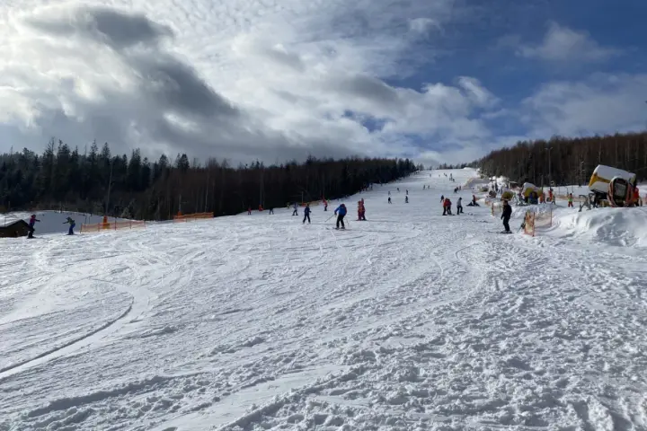 Słoneczny dzień na otwartych stokach narciarskich Wisła. Narciarze i snowboardziści cieszą się jazdą po ośnieżonych trasach, a w tle widać las i maszyny do naśnieżania.