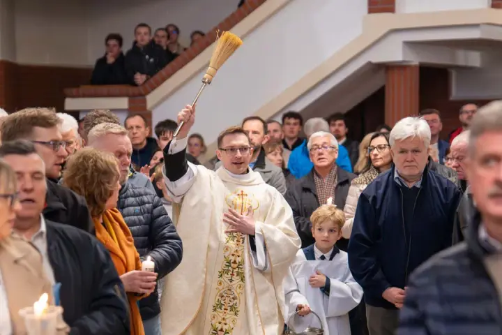 Kapłan błogosławi wiernych podczas liturgii Wielkiej Soboty, unosząc kropidło. W tłumie widać ludzi z zapalonymi świecami.