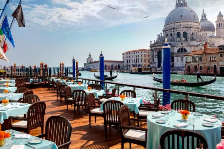 Tavoli apparecchiati con tovaglie azzurre e fiori arancioni, affacciati sul Canal Grande, con gondole e la Basilica di Santa Maria della Salute. Un'ambientazione da sogno per i migliori ristoranti di Venezia.